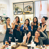 A group of seven people, six standing and one sitting in aprons, smile inside Craft & Culture - Kombucha, Kefir & Probiotics Singapore. They're celebrating their [New & Updated] Kombucha Masterclass Workshop while holding skincare products amid shelves and a poster highlighting fermentation benefits.