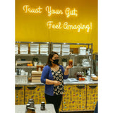 In a kitchen with a bright yellow background and neon sign reading "Trust Your Gut, Feel Amazing," a woman in a blue cardigan and floral top holds a jar, wearing a black face mask. Shelves with jars hint at the [New & Updated] Kombucha Masterclass Workshop by Craft & Culture - Kombucha, Kefir & Probiotics Singapore.