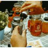 Hands pour liquid from a dark bottle into a clear one, hinting at the [New & Updated] Kombucha Masterclass Workshop by Craft & Culture - Kombucha, Kefir & Probiotics Singapore. Bottles of amber liquid, a metal container, and a potted plant set the scene. A flyer and pen lie partially visible nearby.