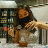 In the New & Updated Kombucha Masterclass Workshop by Craft & Culture - Kombucha, Kefir & Probiotics Singapore, a woman in a black mask carefully pours 'Boulder' into a glass jar with a spoon, surrounded by probiotics-rich shelves.
